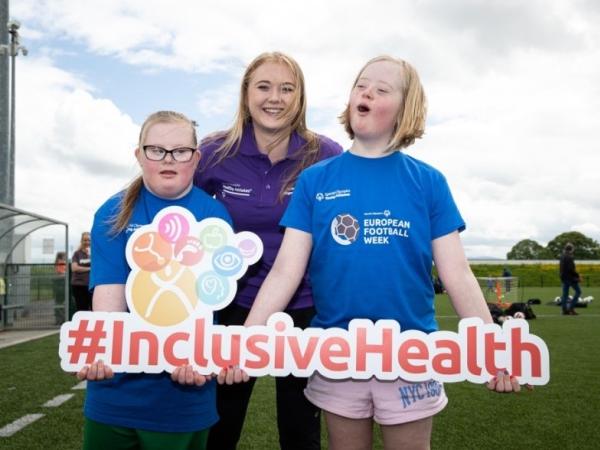 Woman stands with two young athletes holding sign that says inclusive health