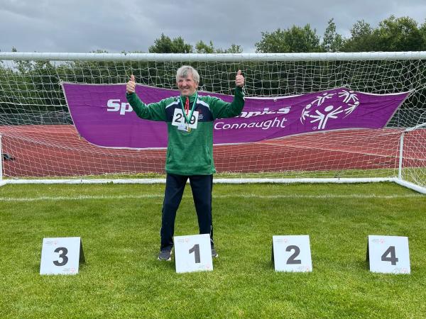 This image shows an Athlete standing in first place with both hands doing a thumbs up in the air and smiling.