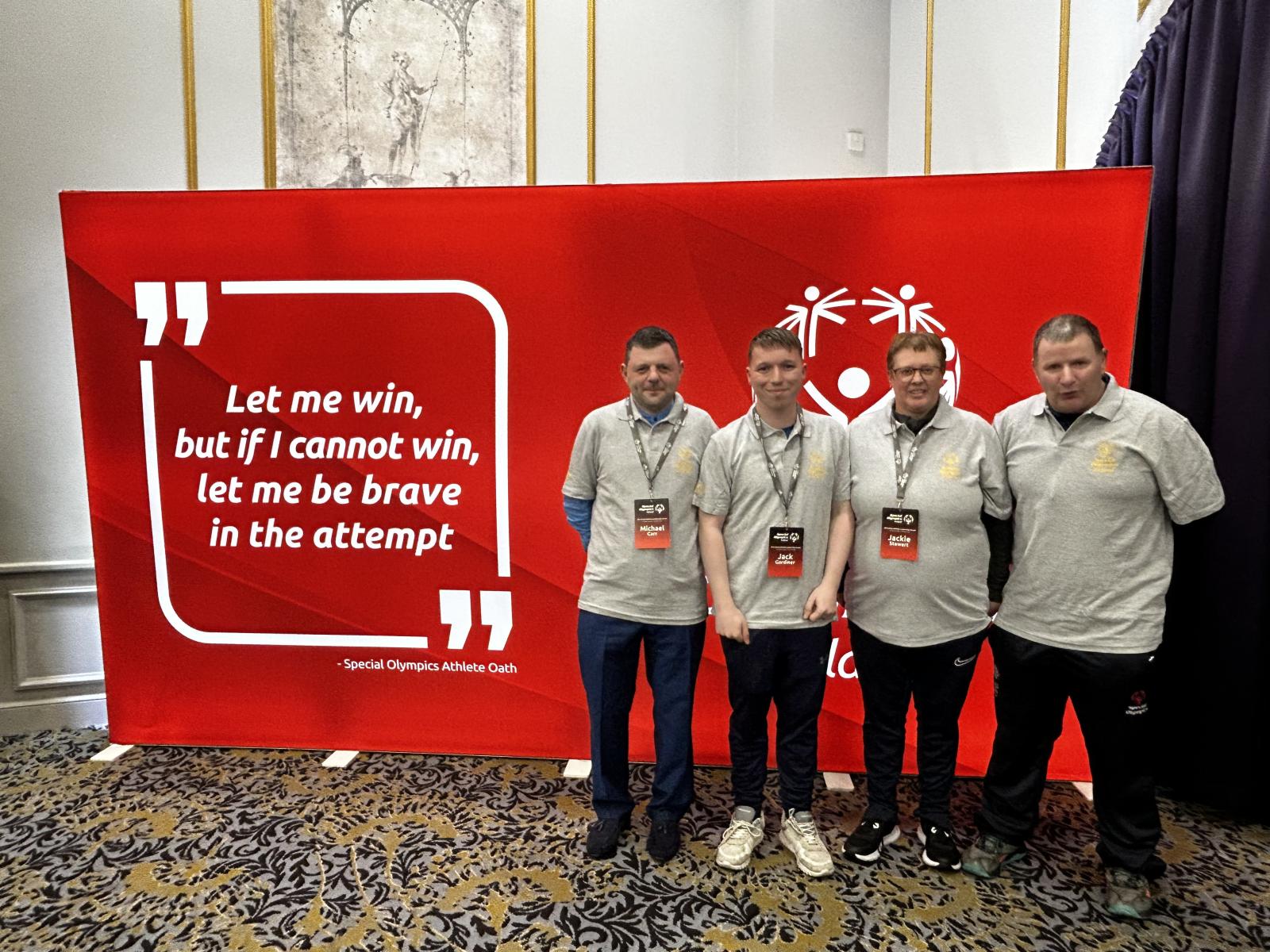 4 members of the Special Olympics Athlete Voices group standing for a photo in front of a large red banner with a quote on it